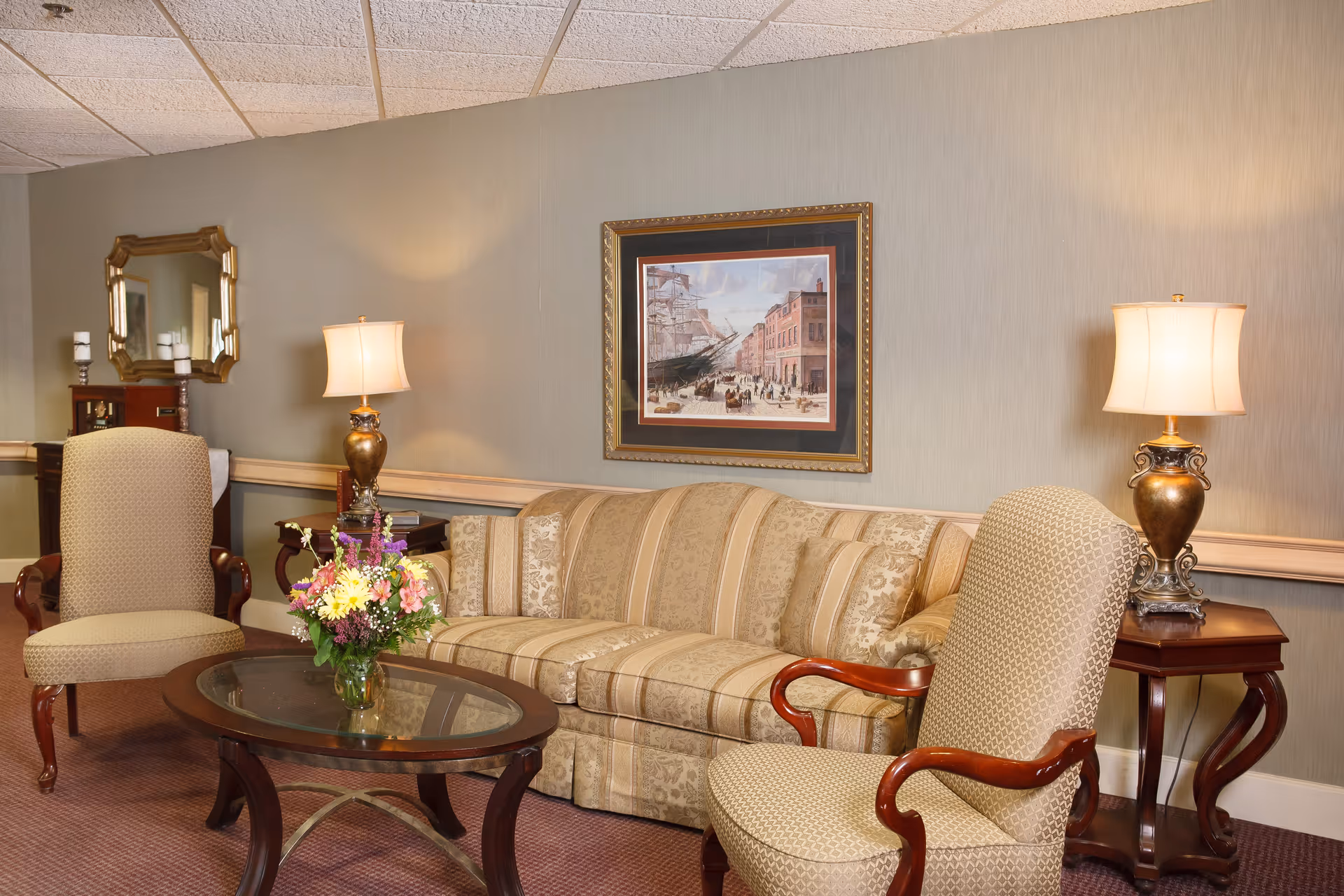 A cozy living room area with a beige patterned sofa and two matching armchairs with wooden armrests. A glass-top coffee table with a flower arrangement is in front of the sofa. Two table lamps with beige shades sit on wooden side tables on either side of the sofa. A framed painting depicting a historical street scene hangs on the wall above the sofa. A mirror and decorative items are visible in the background.