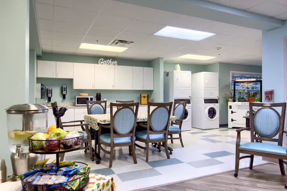 A bright, clean common area in a senior living facility featuring a dining table with six chairs, a kitchen area with white cabinets, a microwave, and two stacked washer and dryer units. A water dispenser with lemon water and a tiered tray of snacks and fruits are visible in the foreground. The word 'Gather' is displayed on top of the cabinets.