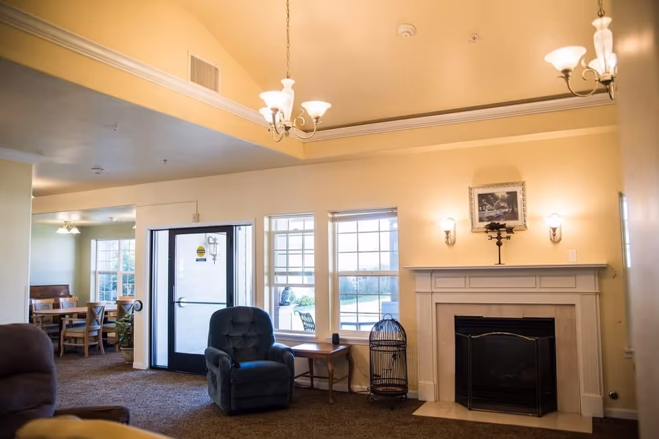 Communal living room with an armchair, fireplace, windows, and a dining area visible through a doorway.