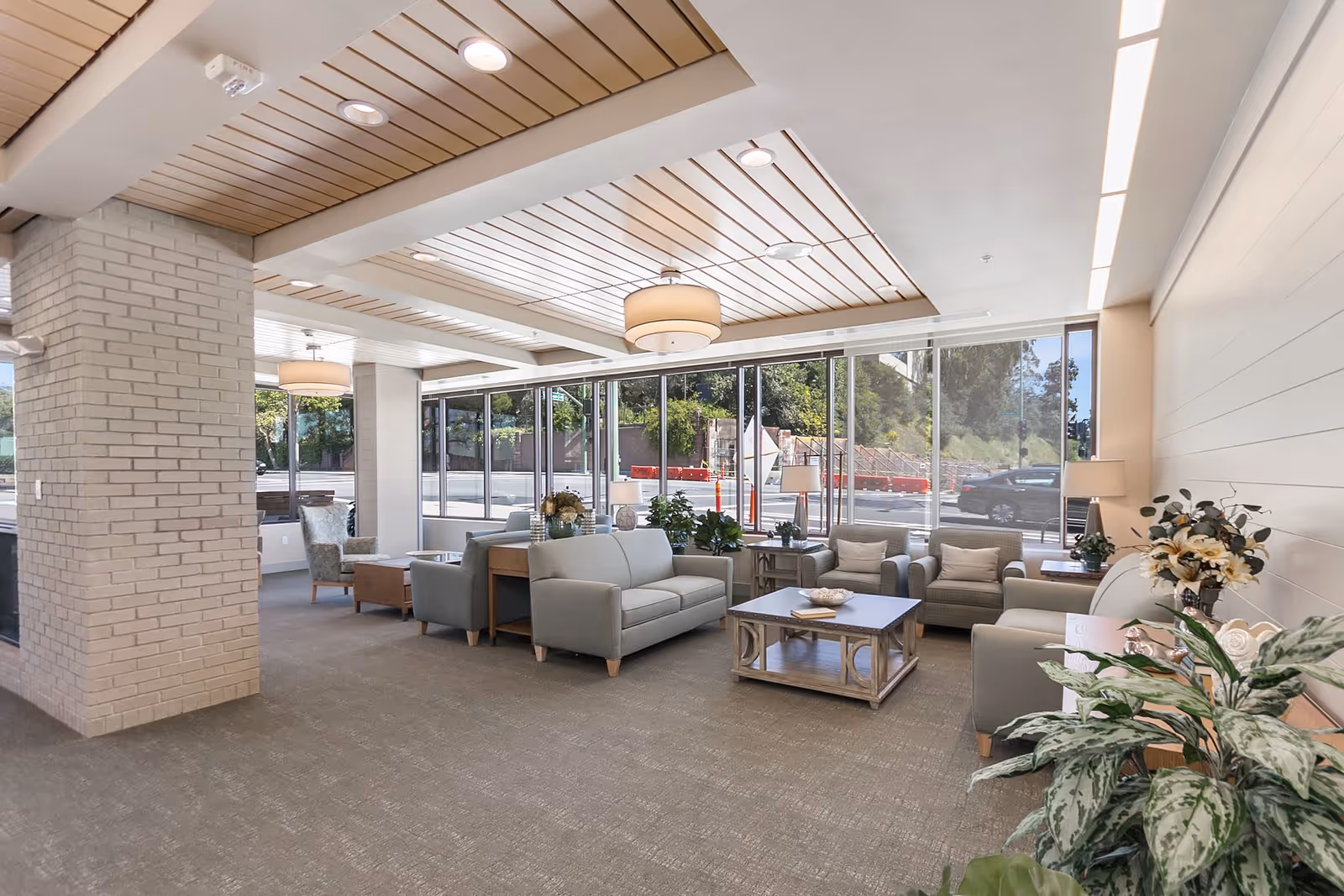 A bright and spacious living room area with large floor-to-ceiling windows allowing natural light to fill the space. The room features light gray sofas and armchairs arranged around wooden coffee tables. There are decorative plants and flowers placed on tables and near the walls. The ceiling has wooden paneling with recessed lighting and modern hanging light fixtures. Outside the windows, a street and greenery are visible.