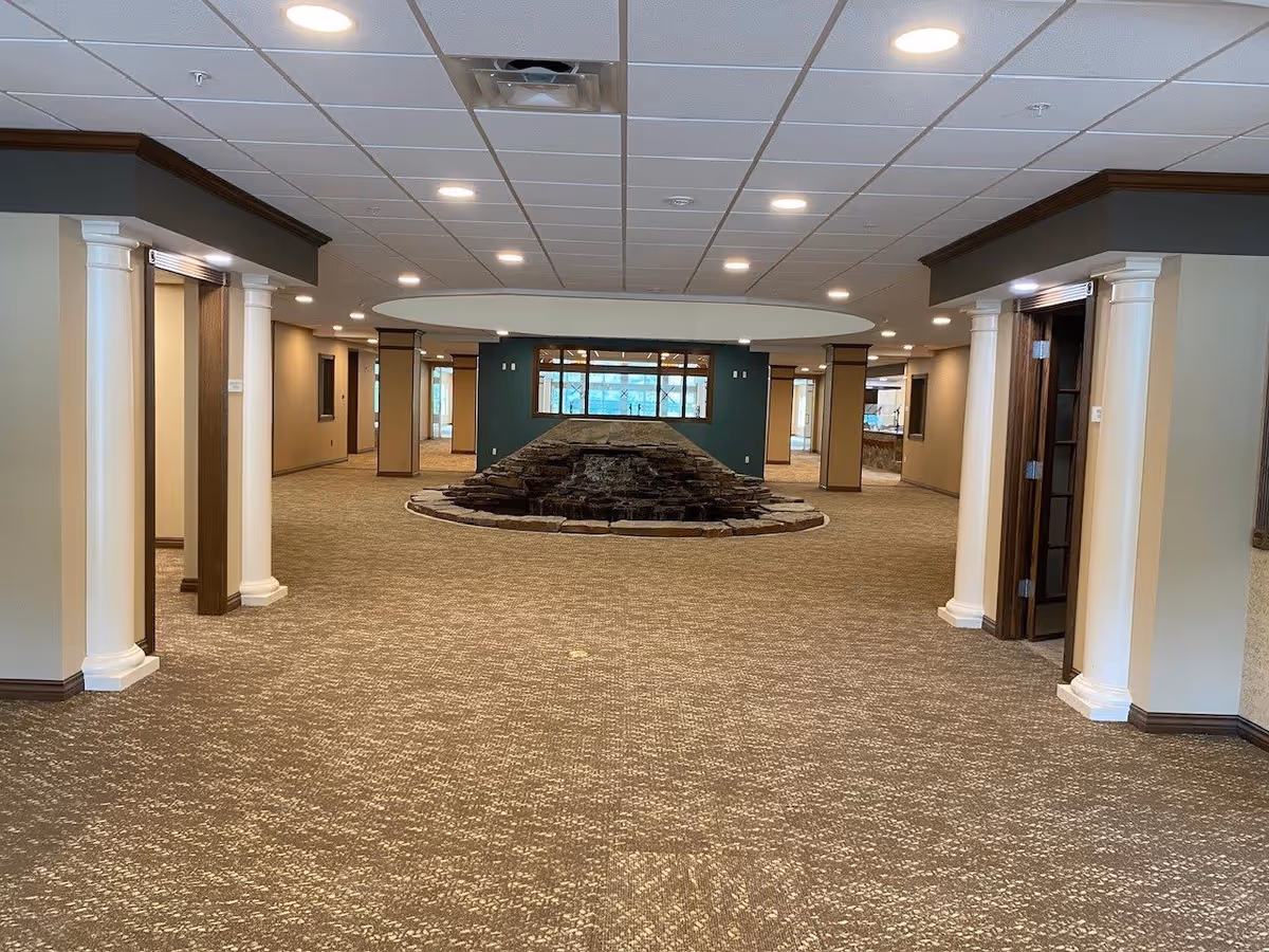 Spacious interior hallway of a retirement facility with beige carpet, white columns, recessed lighting, and a central stone water feature under a circular ceiling design.