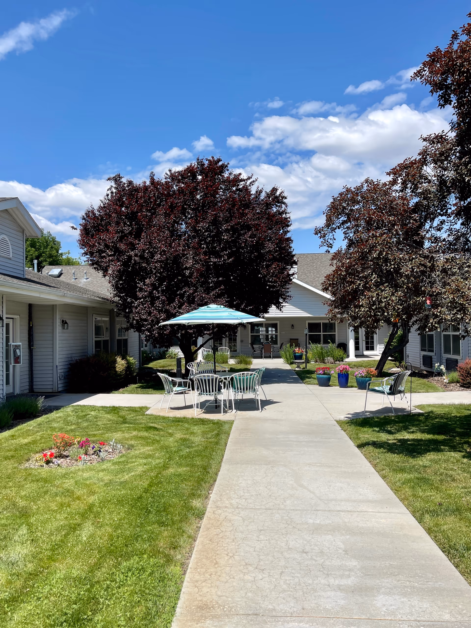 Outdoor courtyard area at Avista Senior Living Garden City featuring a concrete walkway, green grass, flower beds, several trees with dark red leaves, and white metal patio furniture with a blue and white striped umbrella under a bright blue sky with scattered clouds.