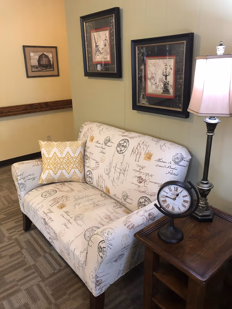 A cozy corner in a room featuring a small upholstered loveseat with script and stamp patterns, a yellow and white patterned throw pillow, a wooden side table with a vintage-style clock and a tall table lamp with a white shade. Two framed pictures hang on the light green wall behind the loveseat.