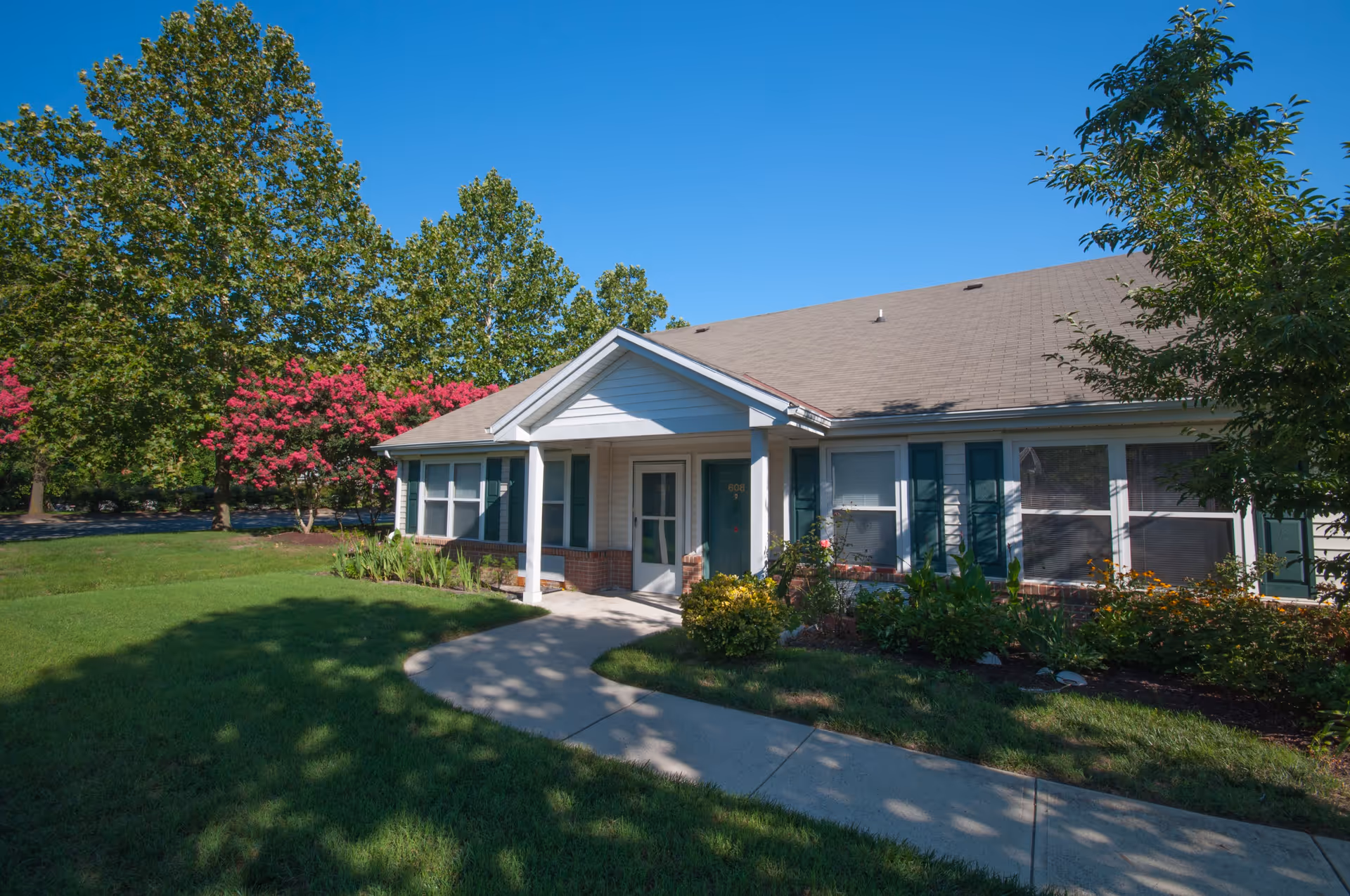 Single-story residential building with white siding and green shutters, surrounded by green grass, trees, and flowering bushes under a clear blue sky.