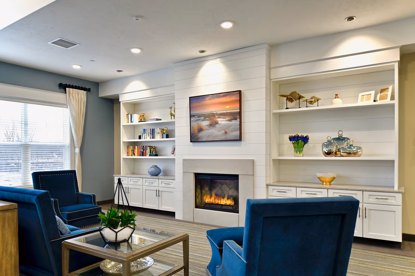 A bright communal living room with blue armchairs, a fireplace flanked by built-in white shelves, and decorative accents.