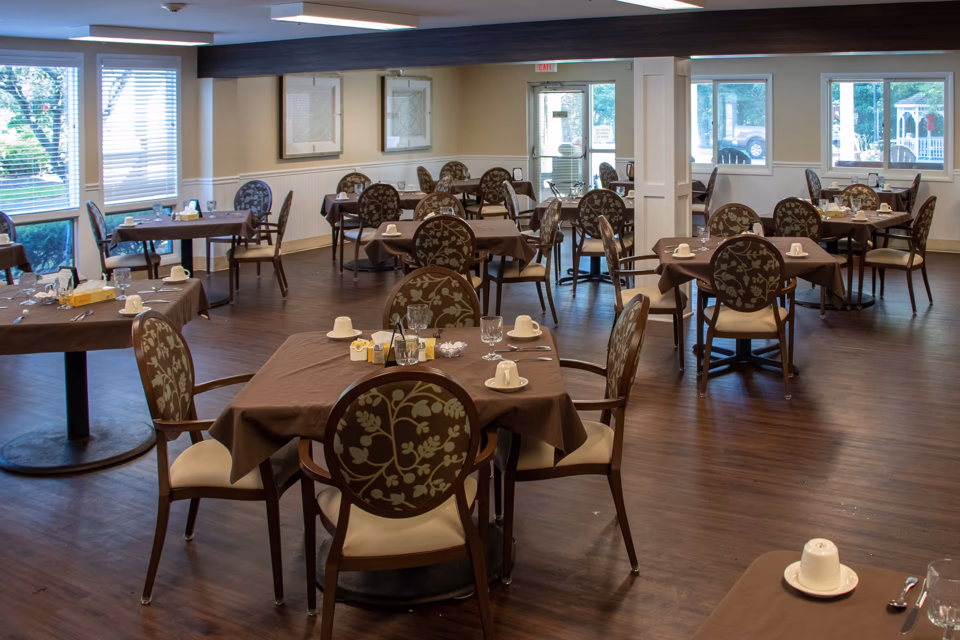 A dining room with multiple tables covered in brown tablecloths, each set with cups, glasses, and utensils. The chairs have floral patterned backs and cream-colored seats. Large windows allow natural light to brighten the room, and framed artwork hangs on the walls.