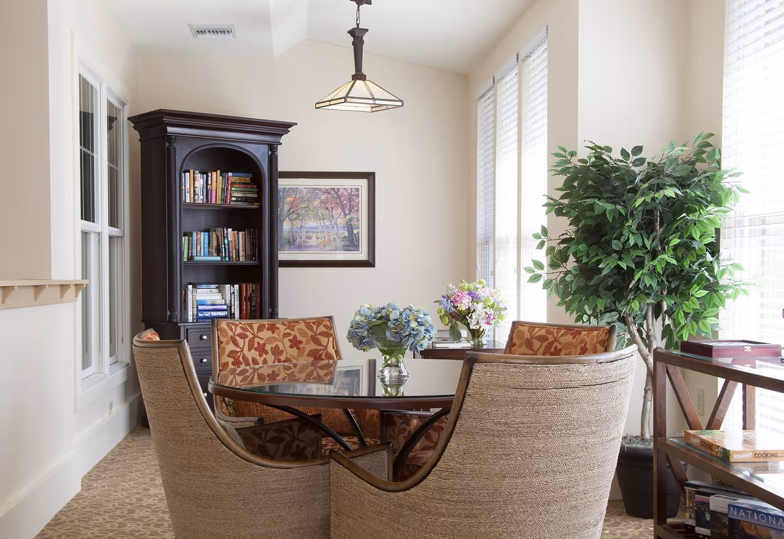 A bright and cozy sitting area with a round glass-top table surrounded by four cushioned chairs, two with floral patterned upholstery and two with plain beige fabric. Behind the table is a tall dark wooden bookshelf filled with books and a framed painting of a forest scene on the wall. To the right, large windows with white blinds allow natural light to fill the room, and a large green potted plant stands next to a wooden side table holding books and a decorative box. A pendant light hangs from the ceiling above the table.