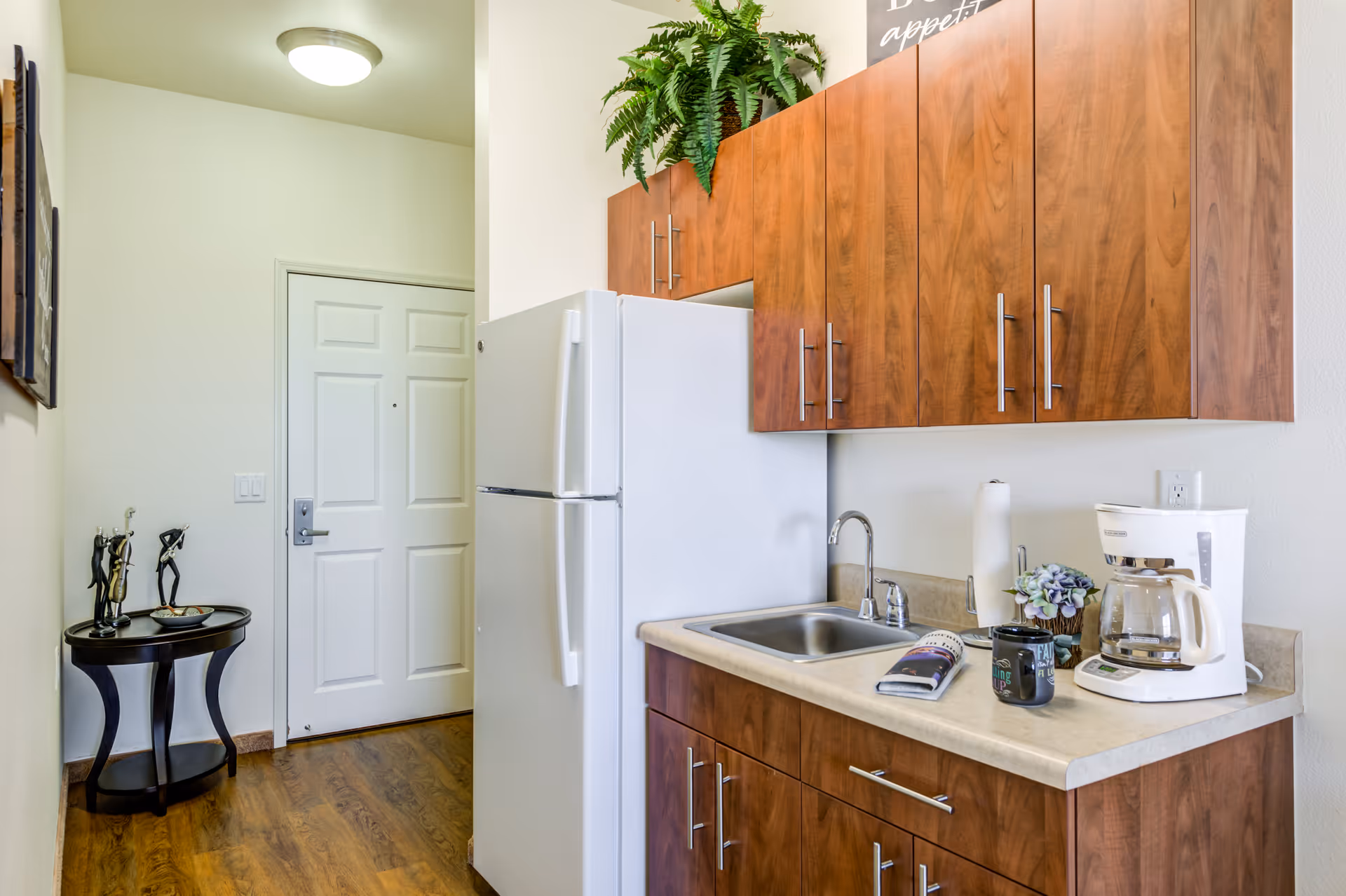 A small kitchen area with wooden cabinets, a white refrigerator, a stainless steel sink, a coffee maker, a paper towel holder, a small plant, and a coffee mug on the countertop. There is a round black side table with decorative items near a white door, and a green plant on top of the upper cabinets.