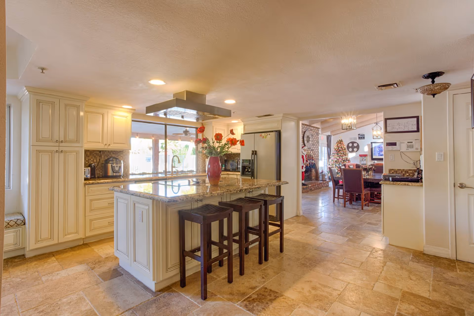 Bright kitchen area with cream-colored cabinets, a large island with a granite countertop, and three dark wooden stools. A red vase with flowers is placed on the island. The kitchen opens into a dining area decorated with a Christmas tree and festive decorations. The floor is tiled with beige stone tiles.