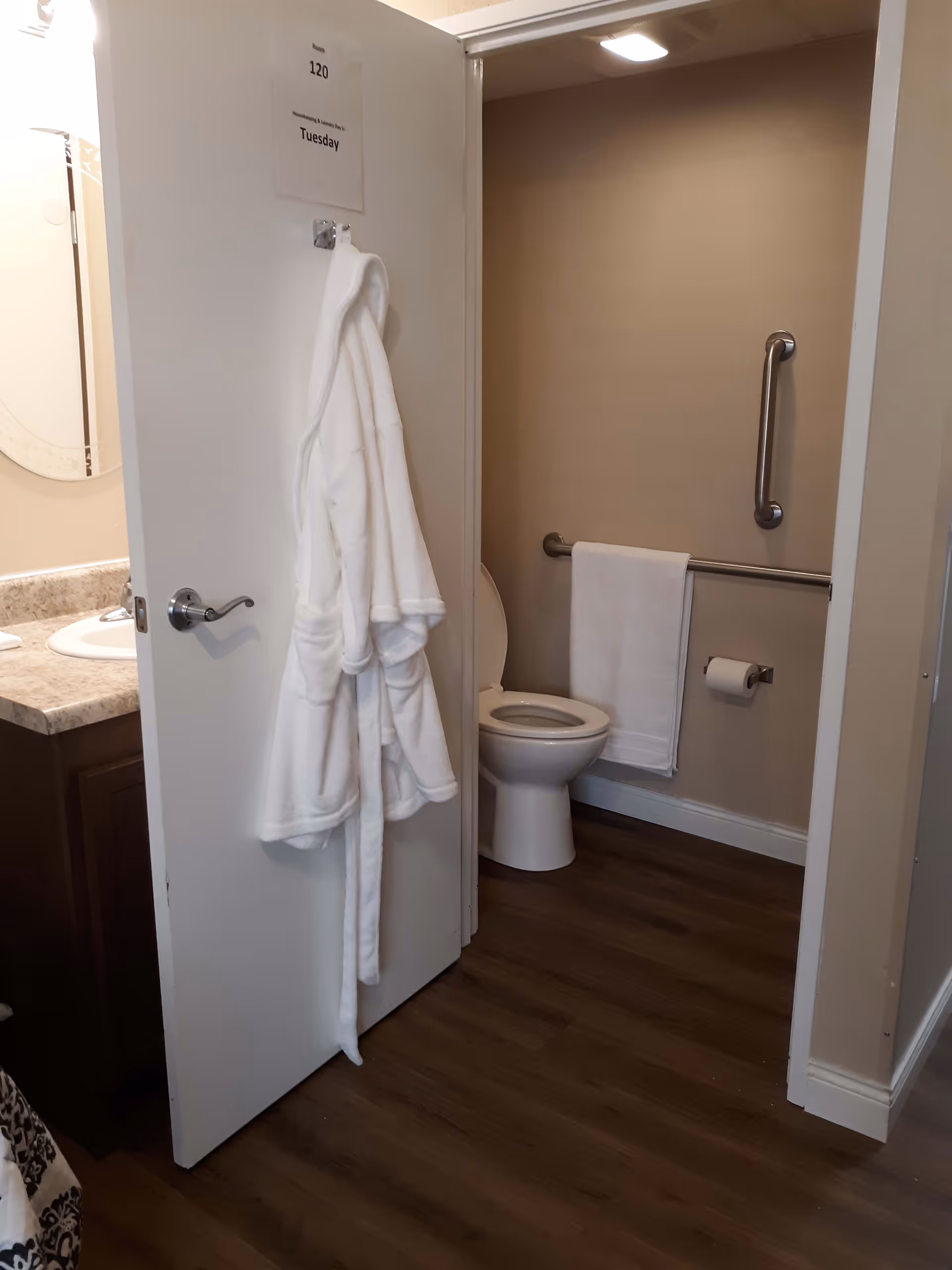 View of a bathroom with an open door showing a white robe hanging on the door. Inside the bathroom, there is a toilet with a grab bar on the wall and a towel hanging on another grab bar. A toilet paper holder is mounted on the wall. Part of a sink and mirror are visible on the left side outside the bathroom.