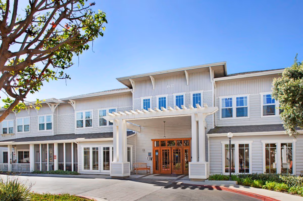 Front exterior of a two-story senior living building with a white pergola-covered entrance, many windows, and landscaped grounds.