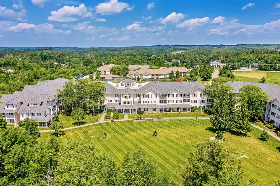 Aerial view of Solstice Senior Living at Bangor showing multiple connected residential buildings surrounded by green lawns, trees, and pathways under a partly cloudy blue sky.