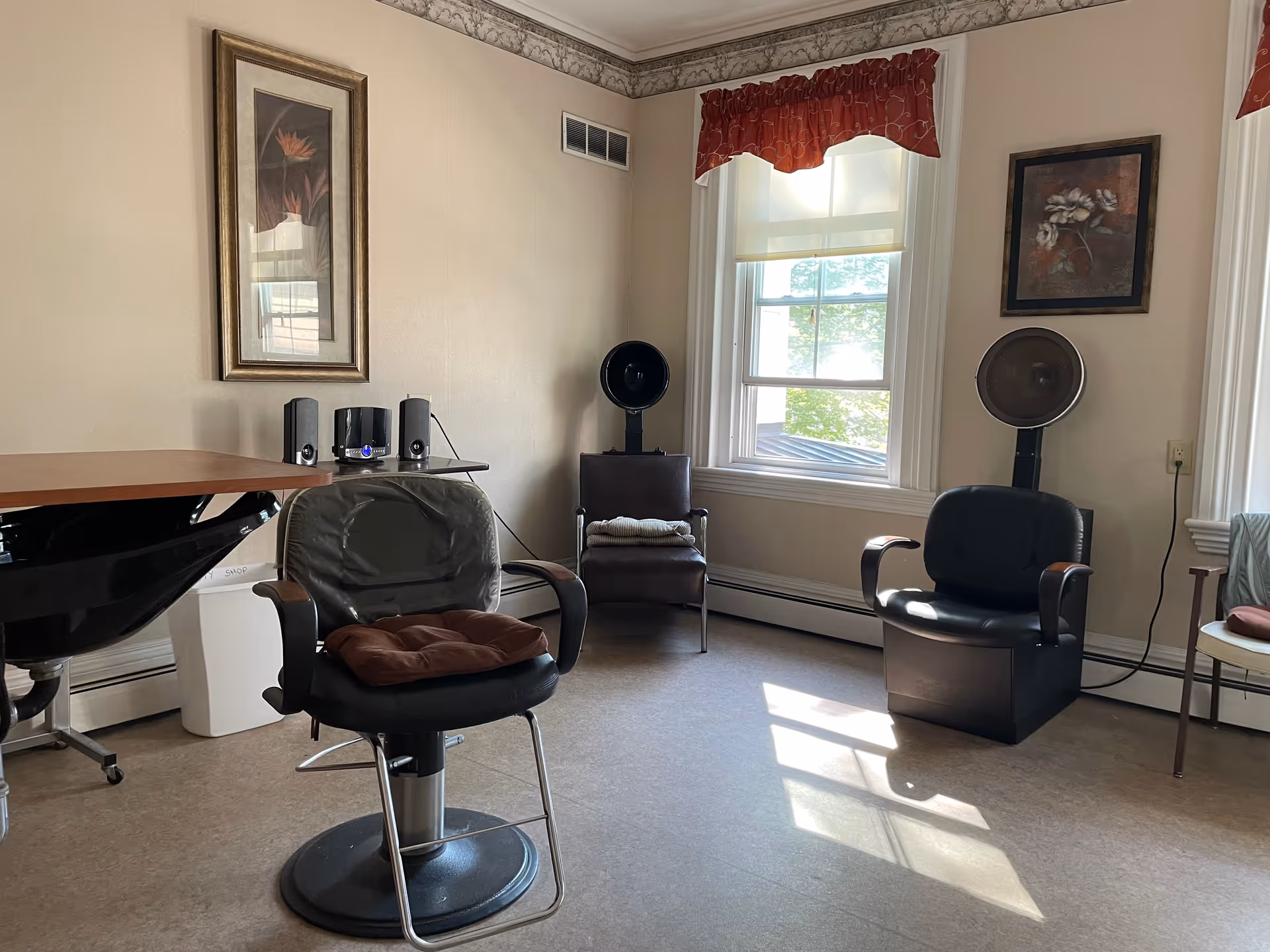 Sunlit interior room with salon chairs, hooded hair dryers, and framed artwork by a window.