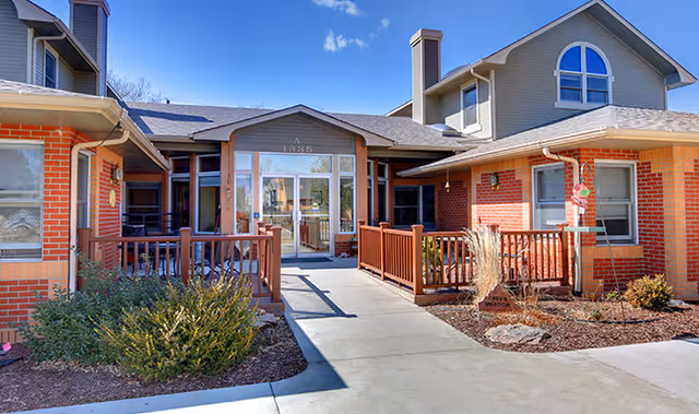 Exterior view of a single-story brick building with a central entrance featuring glass doors and windows. The building has a pitched roof with dormer windows and a wooden railing along the walkway leading to the entrance. There are some shrubs and landscaping on either side of the walkway under a clear blue sky.