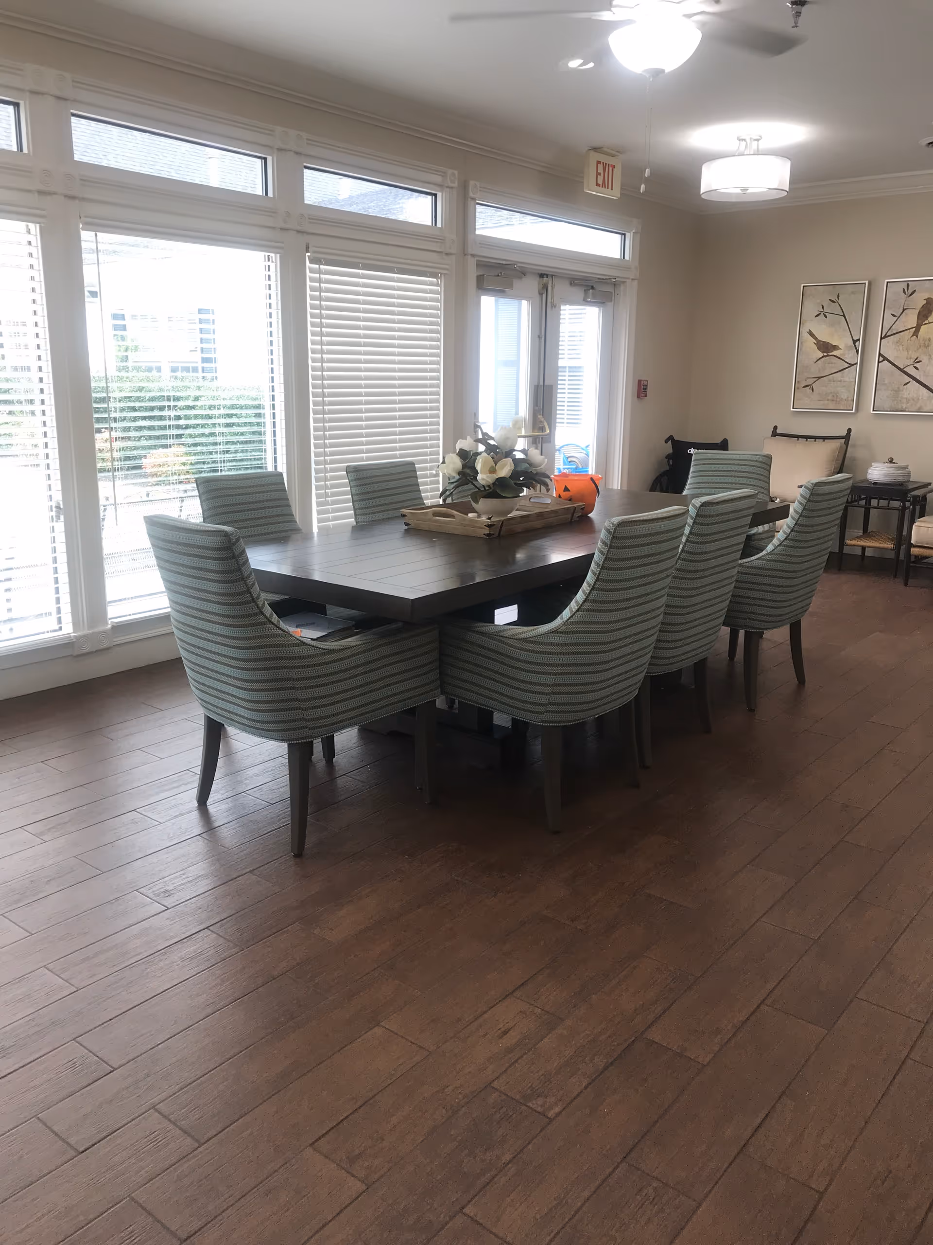 A dining area with a large rectangular wooden table surrounded by eight striped upholstered chairs. The room has large windows with blinds, letting in natural light. There is a floral centerpiece on the table and two framed bird artworks on the wall. The floor is wooden, and there are ceiling lights and an exit sign above a door in the background.