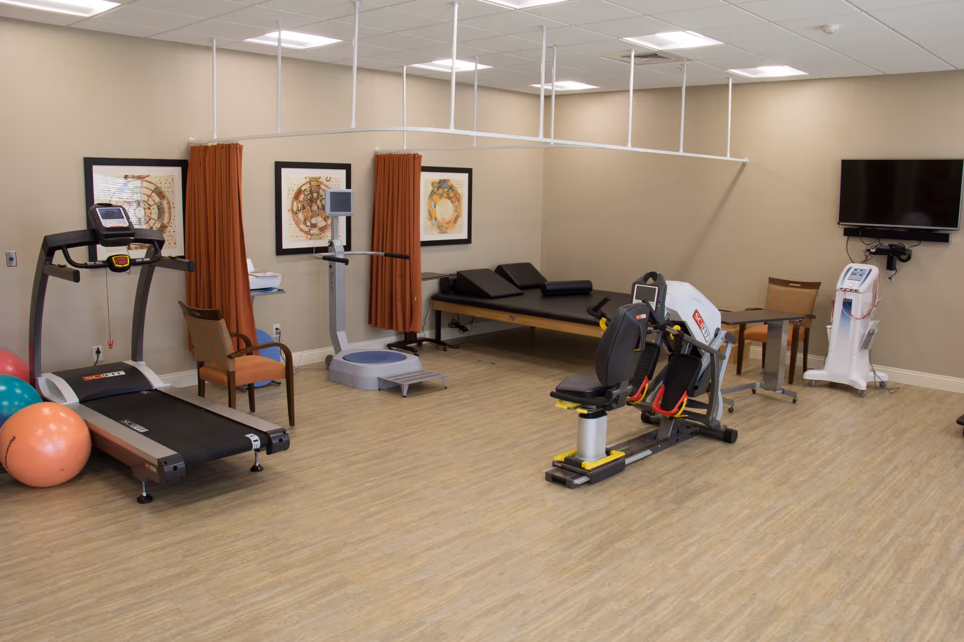 A senior living facility exercise room with a treadmill, exercise bike, balance scale, therapy table, chairs, exercise balls, and a wall-mounted TV. The room has light wood flooring, beige walls, and framed artwork with orange curtains.