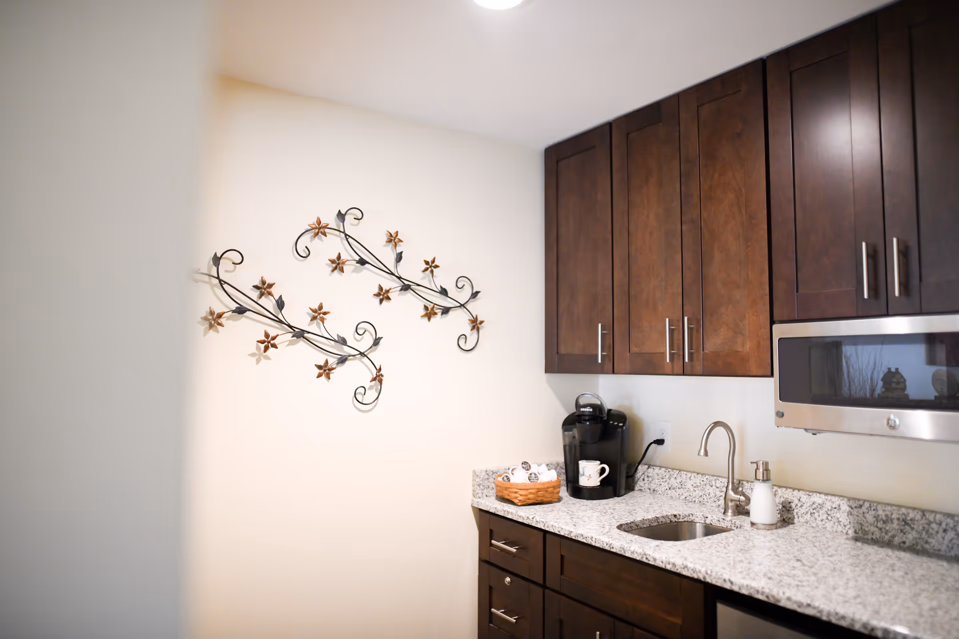 A small kitchen area with dark wooden cabinets, a granite countertop, a stainless steel microwave, a small sink, a coffee maker, and a decorative wall art featuring metal vines with flowers on a light-colored wall.