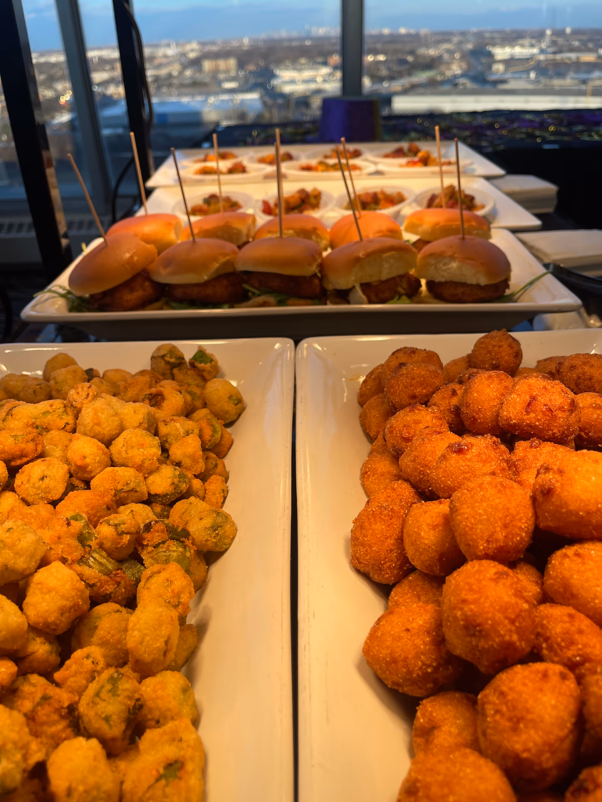 Close-up view of trays filled with fried appetizers including hush puppies and fried okra, with a row of mini sandwiches with toothpicks in the background, set on a table near large windows overlooking a cityscape.