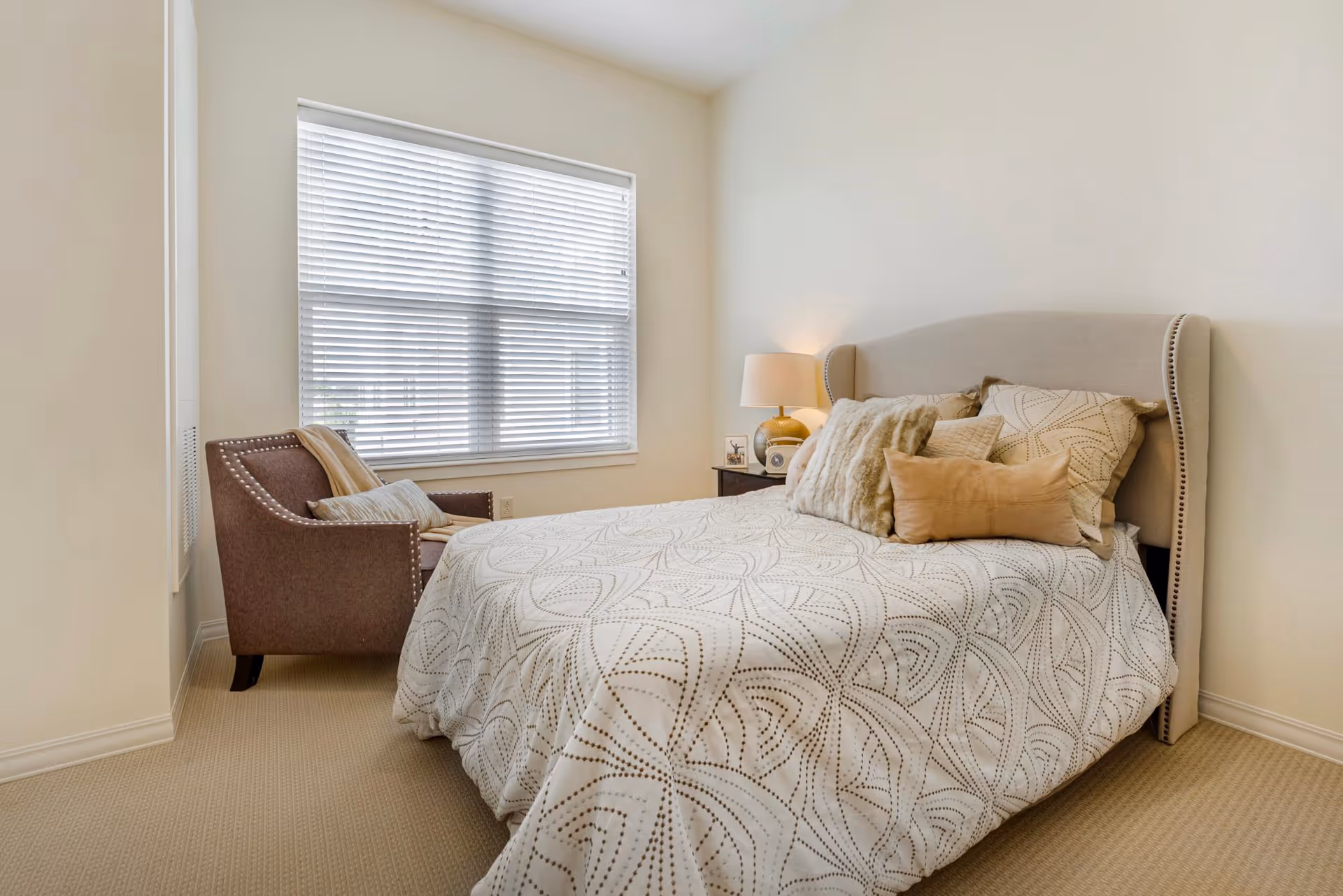 Well-lit bedroom with a bed dressed in patterned bedding, decorative pillows, a nightstand with a lamp, and an armchair by a large window with blinds.