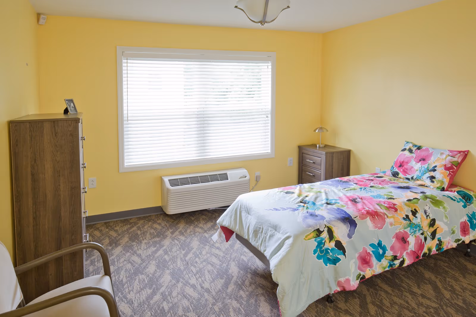 A bright bedroom with yellow walls featuring a single bed with colorful floral bedding, a wooden nightstand with a small lamp, a tall wooden dresser, a window with white blinds, and a beige chair with wooden armrests.