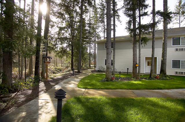 A paved walkway curves through a grassy area with tall trees surrounding it, leading to a two-story beige building with multiple windows and a white door. The scene is outdoors with natural sunlight filtering through the trees.