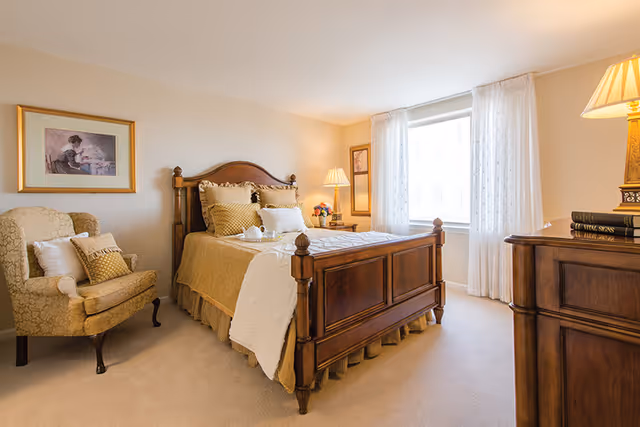 A warmly lit bedroom featuring a wooden bed with beige and gold bedding, a matching armchair with cushions, a wooden dresser with books and a lamp, a window with sheer white curtains, and framed artwork on the walls.