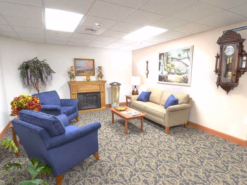 A cozy senior living community lounge with a beige sofa and two blue armchairs arranged around a wooden coffee table. The room features a patterned carpet, a wooden fireplace with decorative items on the mantel, a wall clock, and framed artwork on the walls. There are also plants and a table lamp adding to the warm ambiance.