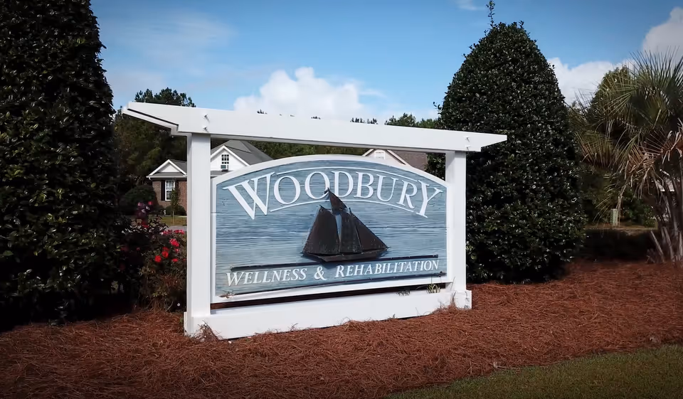 Outdoor sign for Woodbury Wellness & Rehabilitation with a sailboat decoration, surrounded by landscaping including bushes and pine straw mulch, with a house visible in the background under a partly cloudy sky.