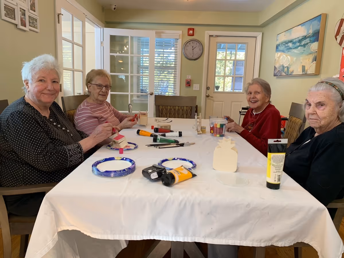 Four elderly women sitting around a table covered with a white tablecloth, engaging in arts and crafts activities with paint, brushes, and wooden shapes in a well-lit room with glass doors and a clock on the wall.