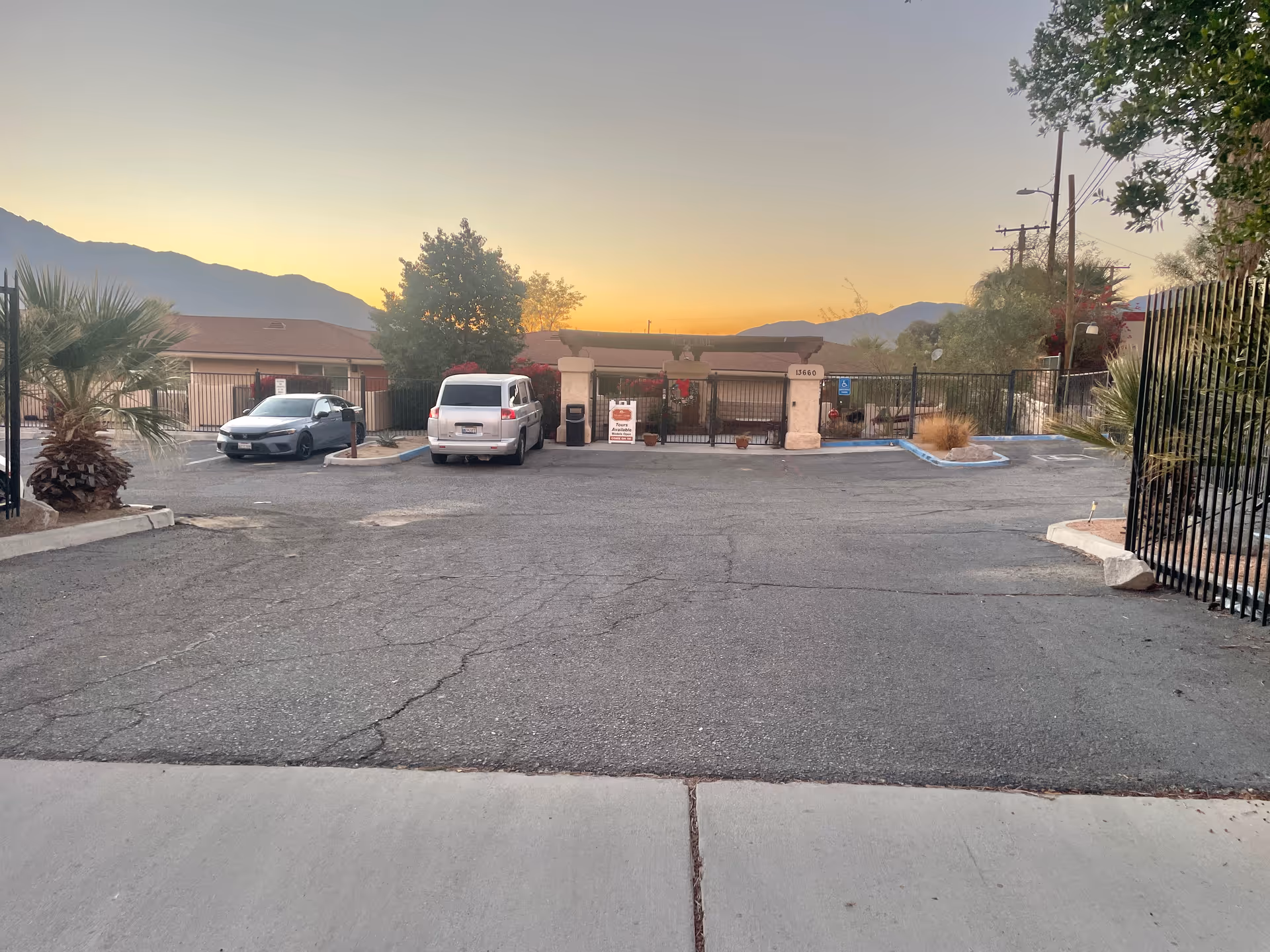Parking lot of Desert Cove Assisted Living At Desert Hot Springs during sunset, showing two parked cars, a gated entrance with a sign, trees, and mountains in the background.