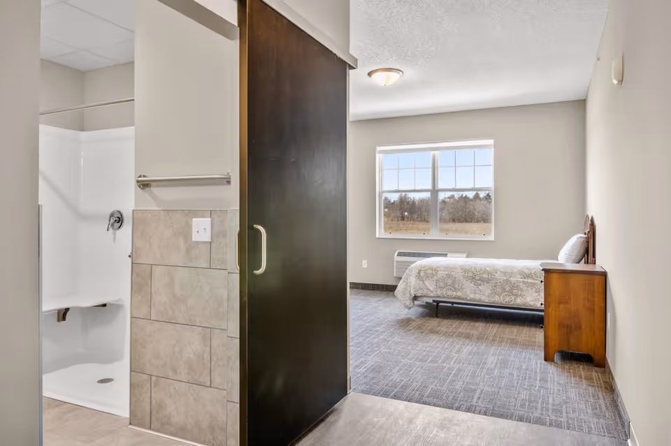 Interior view of a senior living bedroom with a single bed and nightstand and an adjacent bathroom with a walk-in shower separated by a dark sliding door.