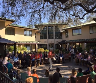 A large group of elderly people seated in a courtyard area of a senior living facility, listening to a person standing and speaking in the center. The courtyard is surrounded by two-story buildings with windows and shaded walkways. Trees provide some shade over the area.