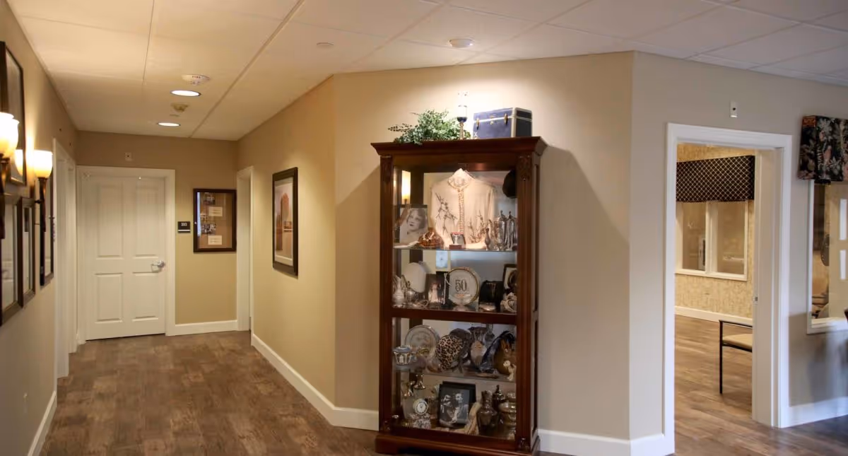 Interior hallway of a senior living facility with beige walls, wood flooring, and framed pictures on the left wall. A glass display cabinet filled with various decorative items and memorabilia is positioned on the right side near an open doorway leading to another room with a chair and window.