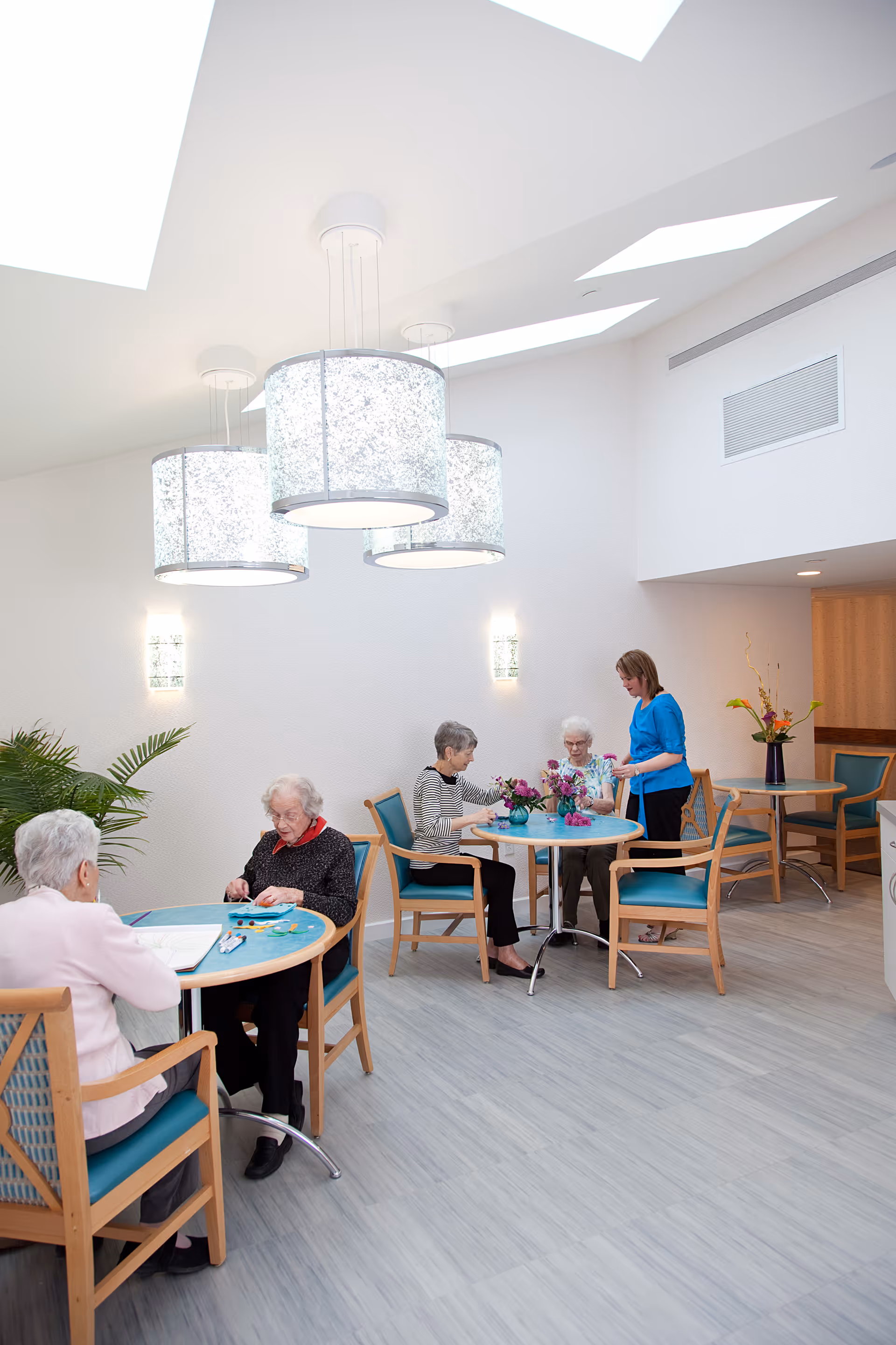 Elderly residents seated at round tables in a bright communal dining/activity room with hanging drum pendant lights.