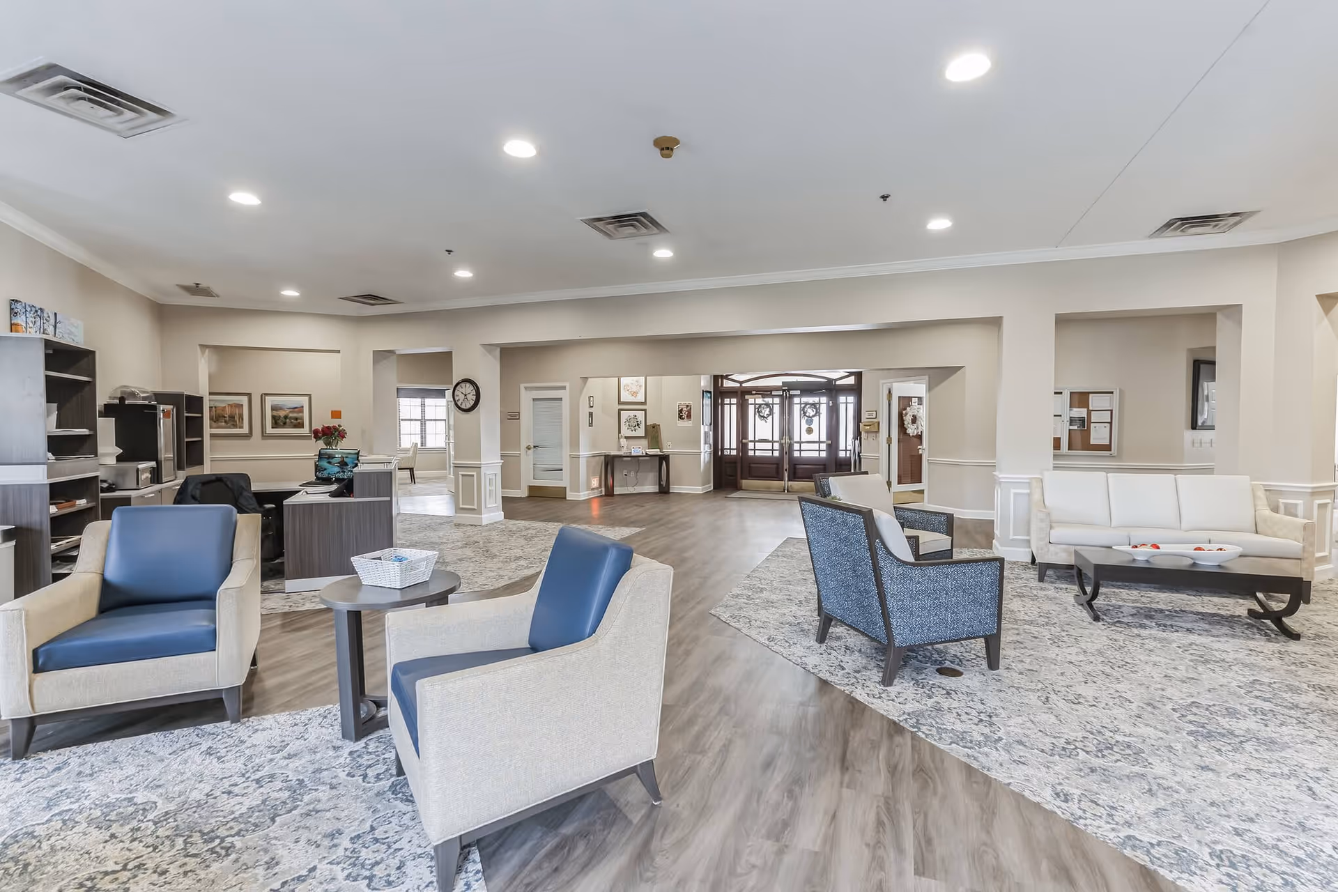 A spacious and well-lit senior living facility common area with multiple seating arrangements including armchairs and sofas on patterned rugs. The room has light-colored walls, wood flooring, recessed ceiling lights, and a reception desk area with office equipment. Large windows and glass doors are visible in the background.
