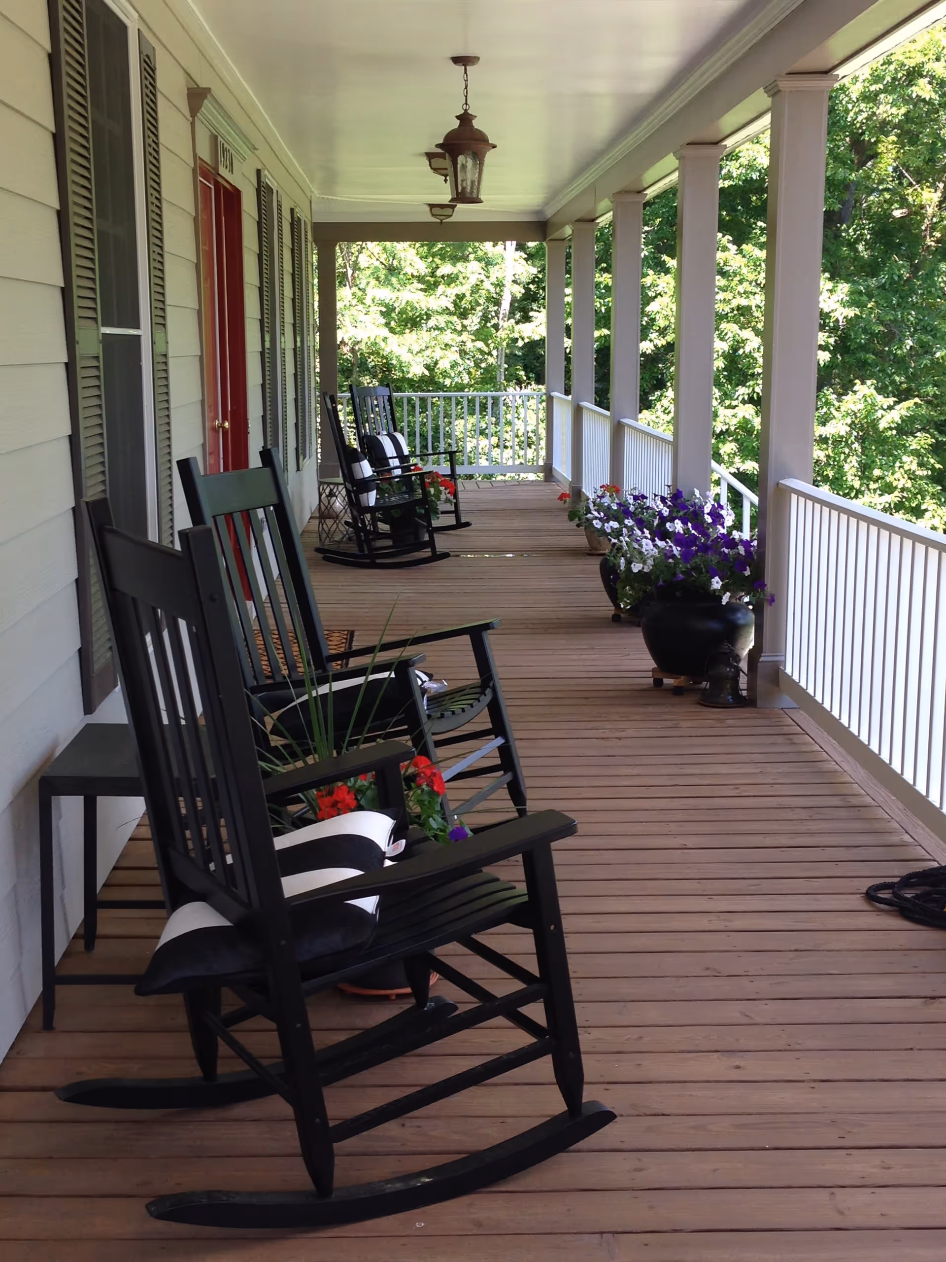 A covered wooden porch with several black rocking chairs, each with a black and white cushion. There are potted flowers placed along the porch railing, and the porch overlooks a green, leafy outdoor area. The porch has white railings and columns, and a red door is visible on the left side.