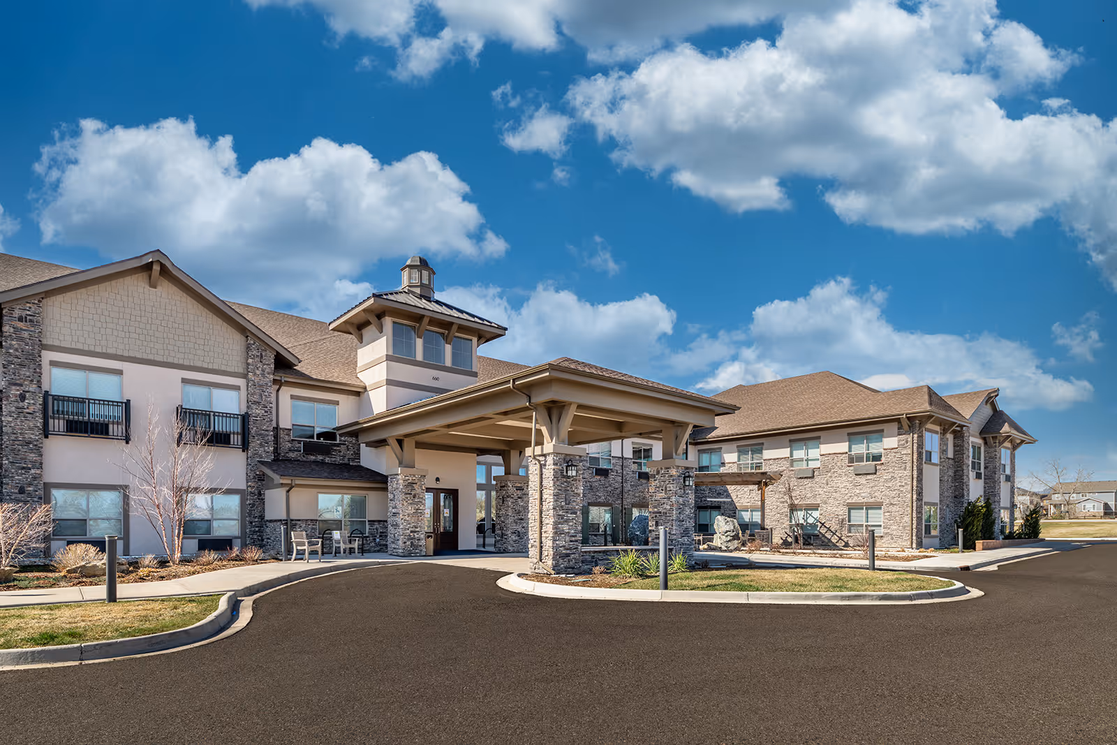 Exterior view of The Peaks at Old Laramie Trail senior living facility showing a large two-story building with stone and beige siding, a covered entrance with stone pillars, and a clear blue sky with scattered clouds.