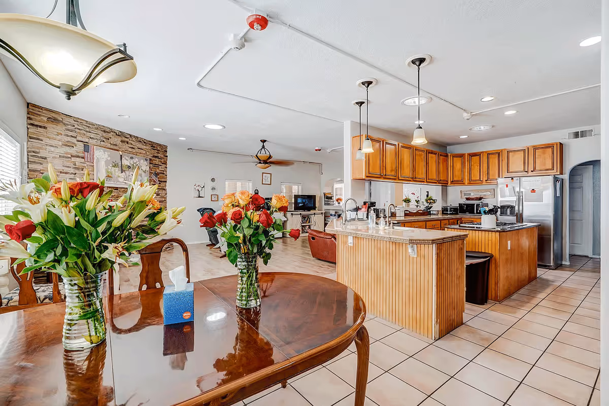 A bright and spacious open-concept kitchen and living area in a senior living facility. The kitchen features wooden cabinets, a large island with a sink, and stainless steel appliances. In the foreground, a polished wooden dining table holds two vases with colorful flowers and a box of tissues. The living area in the background has comfortable seating and a TV, with a stone accent wall decorated with framed pictures and small American flags.