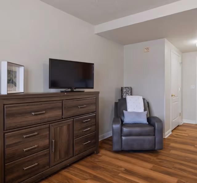 Interior view of a room with a wooden dresser topped by a flat-screen TV on the left and a dark gray armchair with a white blanket and a blue pillow on the right, set against light-colored walls and wood flooring.