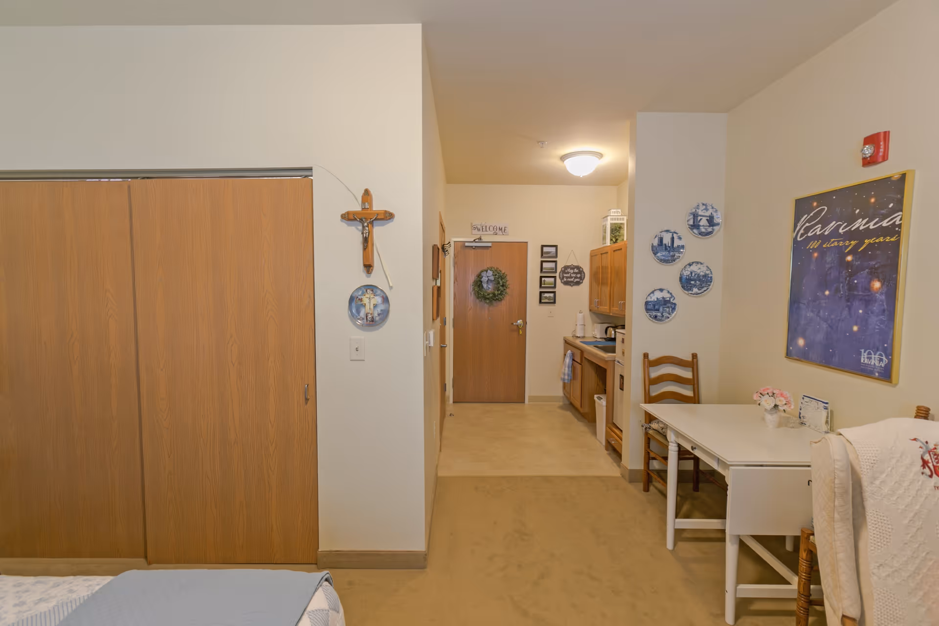 Interior view of a senior living facility room at Heritage Woods of Gurnee showing a bed partially visible on the left, a wooden closet with sliding doors, a small kitchenette area with cabinets, a sink, and a microwave, a white table with a wooden chair, and decorative items on the walls including a crucifix, decorative plates, and framed artwork.