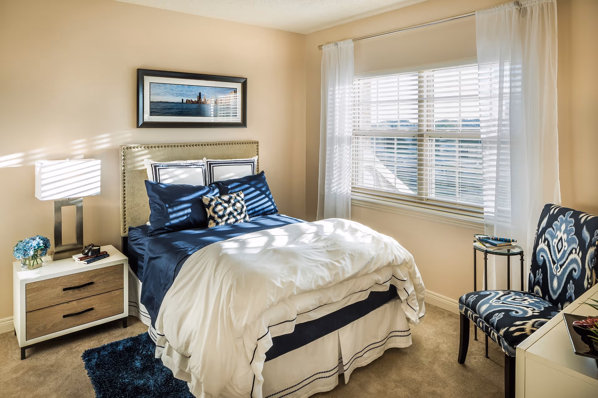 A bright bedroom with a neatly made bed featuring navy blue and white bedding. There is a nightstand with a modern lamp and a small vase of blue flowers to the left of the bed. A framed picture hangs above the bed. To the right, there is a window with white blinds and sheer curtains, and a patterned blue and white chair next to a small round table.