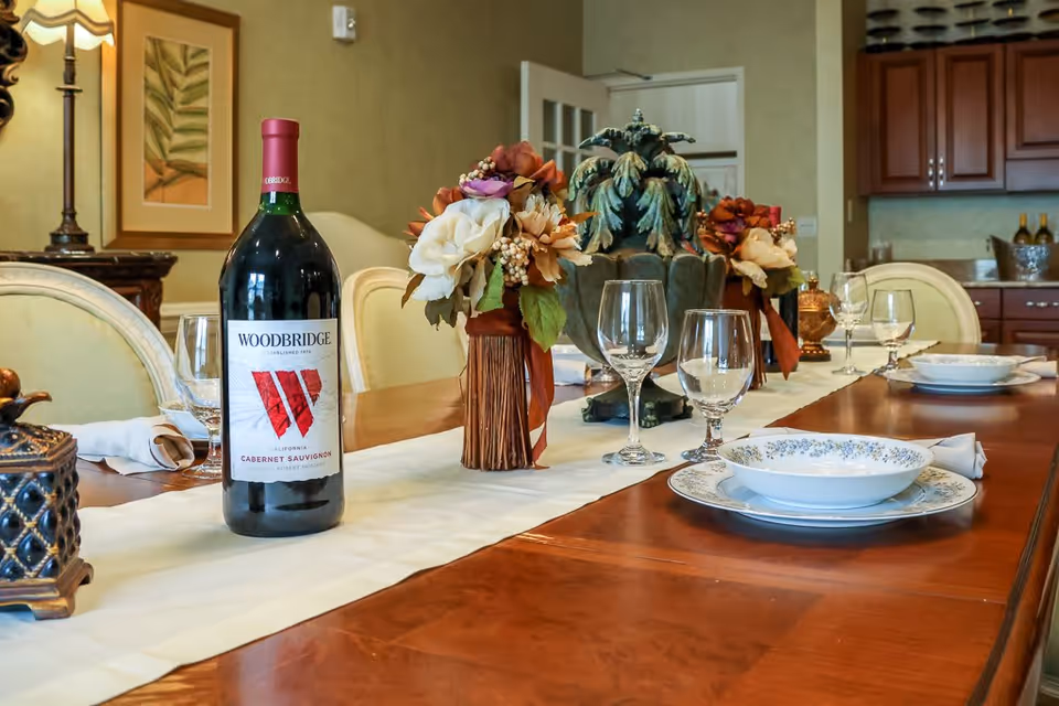 A dining table set with a large bottle of Woodbridge Cabernet Sauvignon wine, floral centerpieces, wine glasses, and plates with bowls. The room has wooden cabinetry and a lamp in the background.
