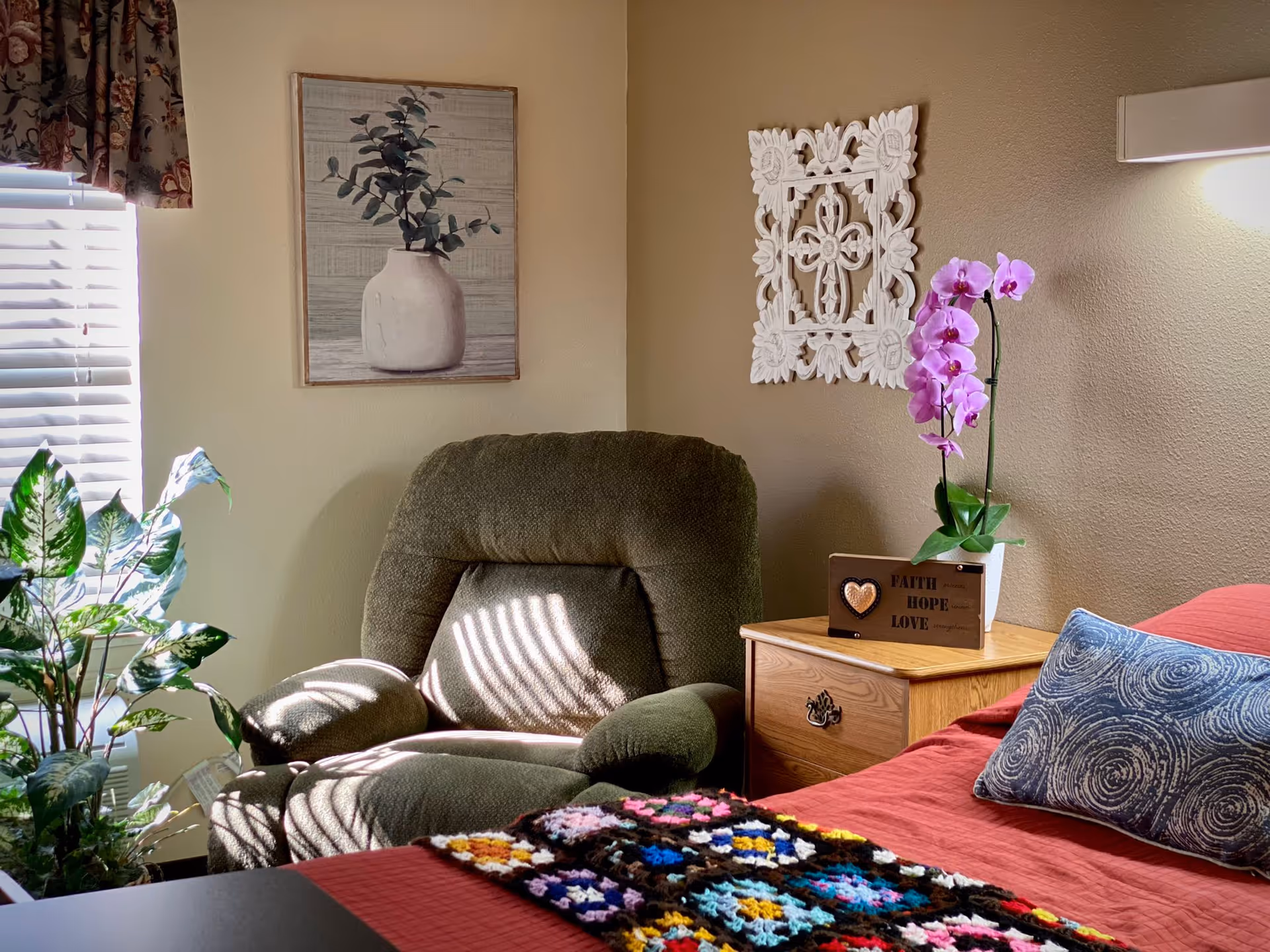 A cozy bedroom corner with a green armchair next to a wooden nightstand. The nightstand holds a potted orchid with pink flowers and a decorative block with the words 'Faith Hope Love'. The bed has a red bedspread, a blue patterned pillow, and a colorful crocheted blanket. The wall features a framed picture of a vase with leaves and a white decorative wall hanging. A window with white blinds and floral curtains lets in natural light, and a green leafy plant is placed near the window.