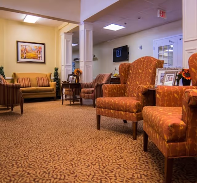 A cozy living room area in an assisted living facility featuring patterned carpet, several upholstered armchairs with orange and red leaf designs, a striped sofa, a yellow couch with striped cushions, a side table with framed photos and flowers, and a framed landscape painting on the wall.