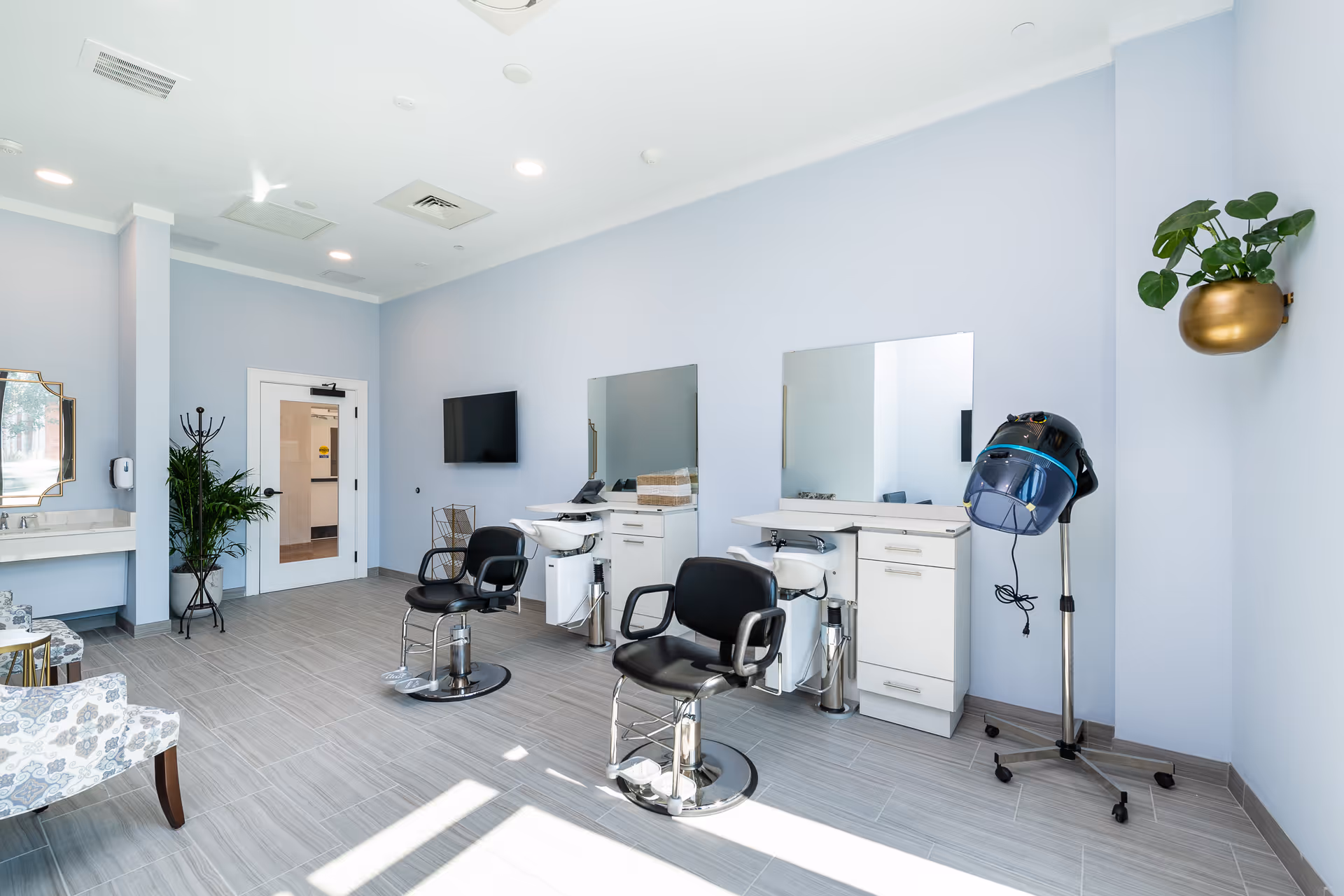 A bright and clean hair salon area inside a senior living facility with two black salon chairs in front of mirrors and wash basins. There is a hair dryer on a stand to the right, a coat rack with a plant near the door, and a TV mounted on the wall. The room has light blue walls and large windows letting in natural light.