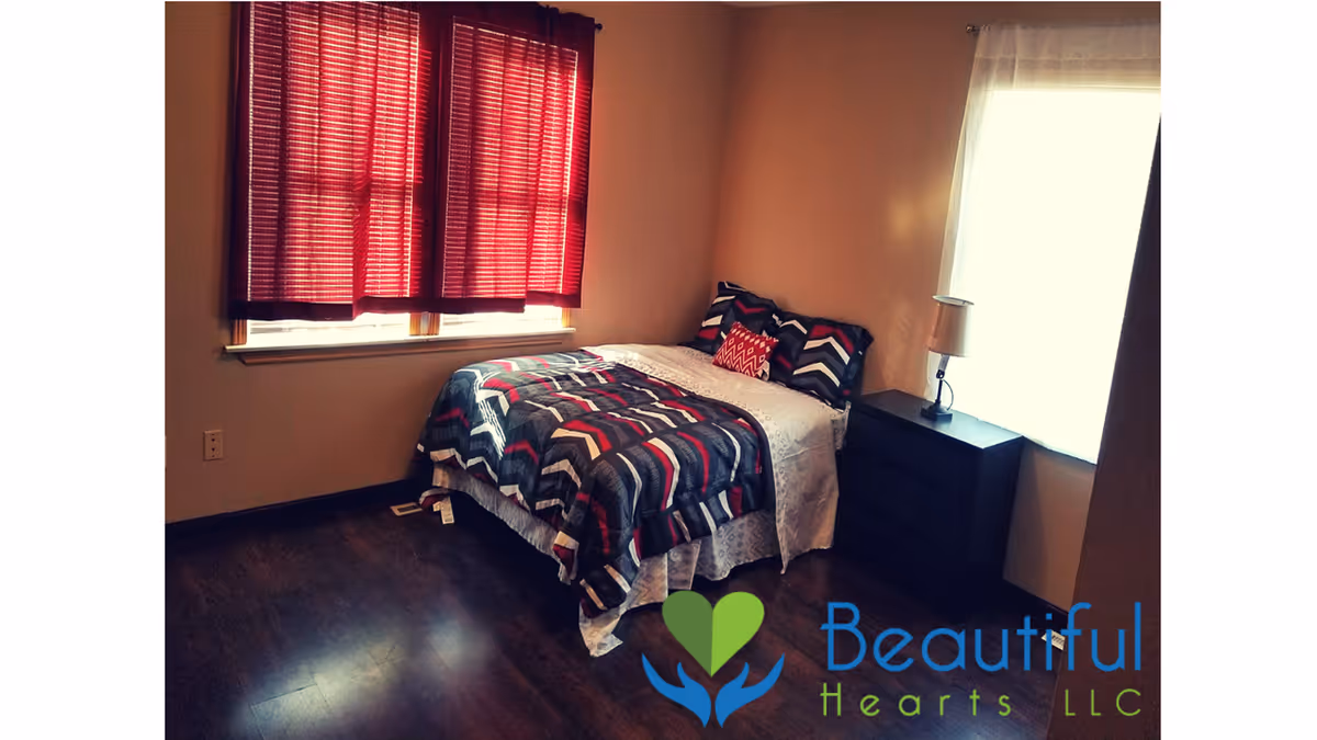 A bedroom with a bed covered in a patterned comforter and pillows, a black nightstand with a lamp, two windows with red and white curtains, and dark wood flooring.