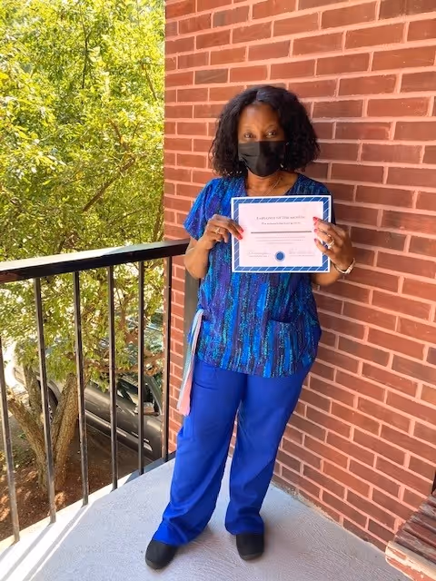 A woman wearing blue scrubs and a black face mask stands on a balcony next to a brick wall, holding a certificate. There are green trees and a parked car visible in the background.