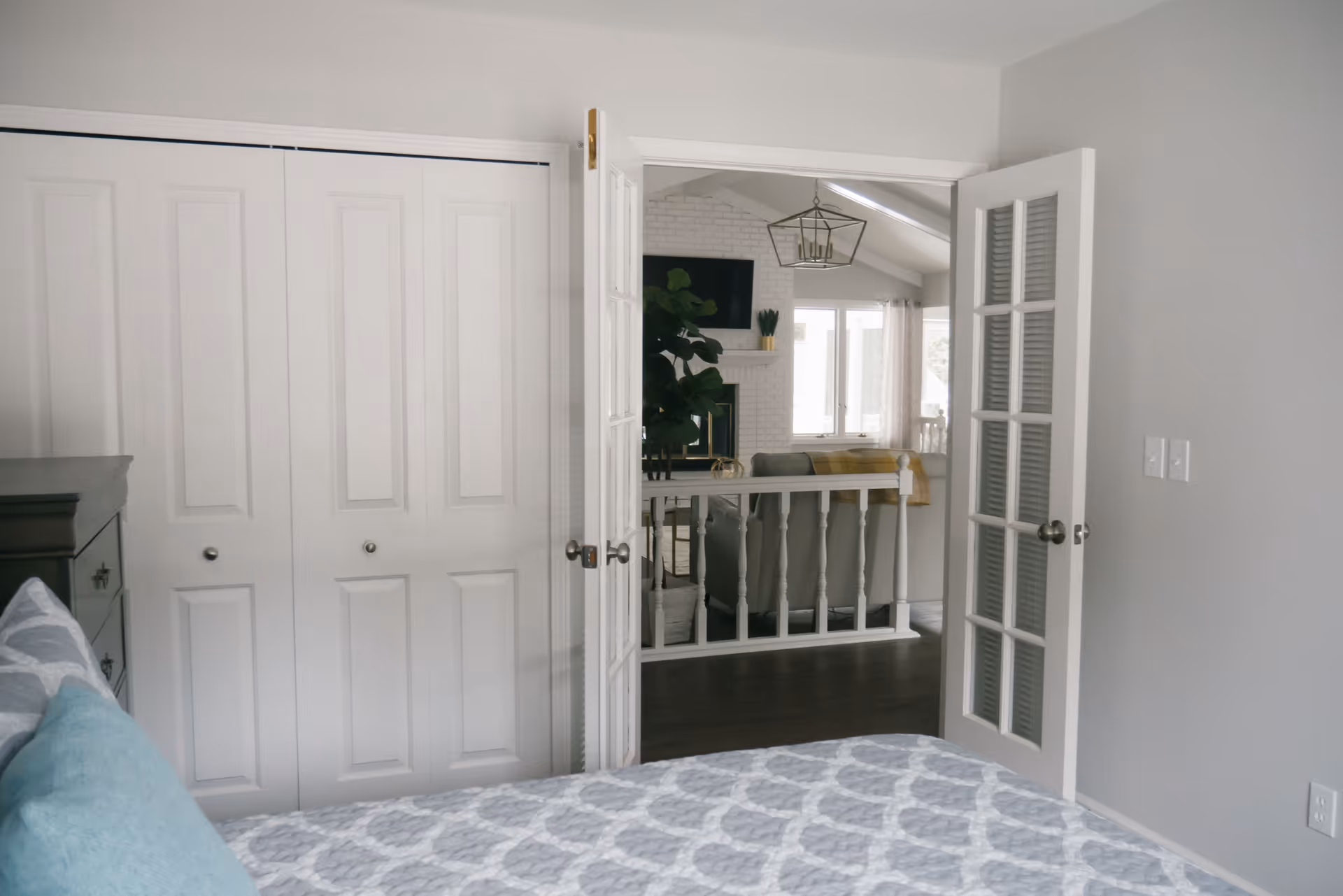 A light gray bedroom with a patterned bed in the foreground, closet doors on the left, and open French doors revealing a bright living room beyond.