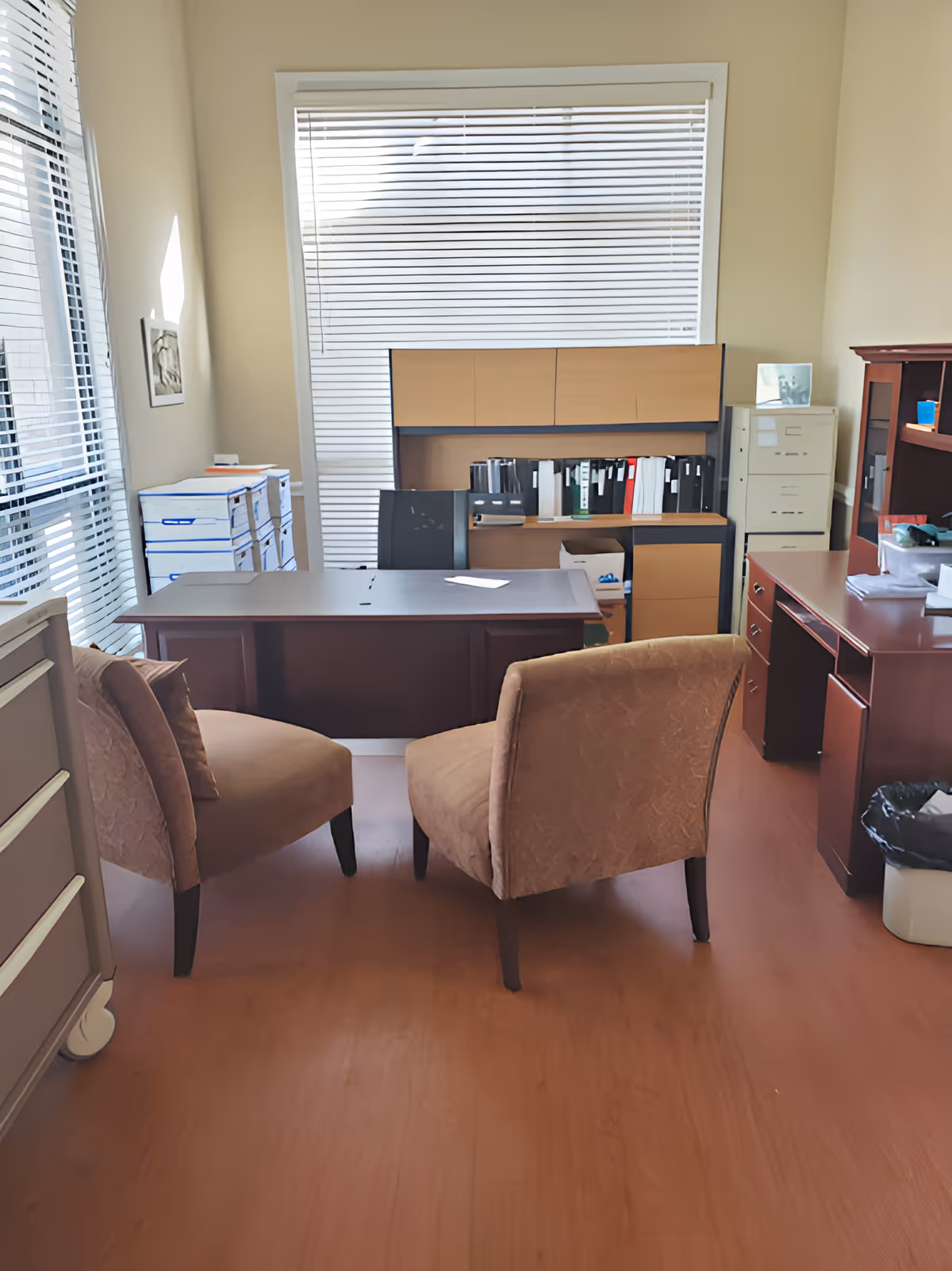 An office room with two upholstered chairs facing a large wooden desk. Behind the desk is a black office chair and a wooden cabinet with shelves holding binders and files. To the right, there is a filing cabinet and another wooden desk with office supplies. The room has large windows with blinds allowing natural light to enter.