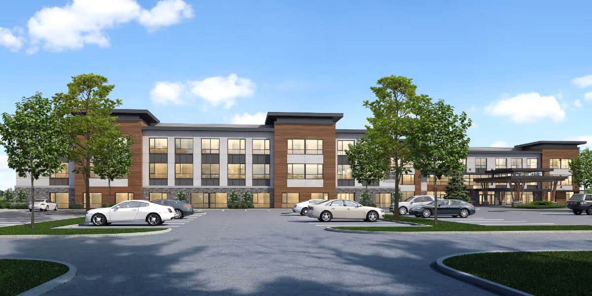 Front exterior view of a modern three-story senior living facility building with large windows, wood and stone accents, surrounded by a parking lot with several cars and landscaped areas with trees and grass under a blue sky with some clouds.